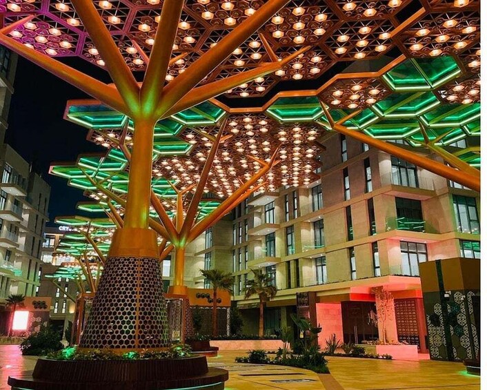 A brightly lit outdoor area with a large, illuminated tree-like structure and modern buildings in the background. 4