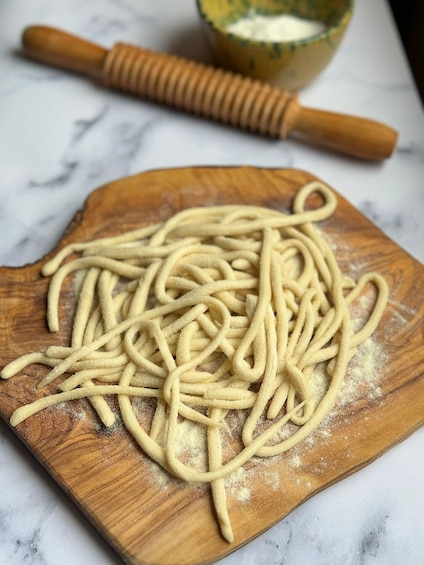 Uncooked spaghetti on a wooden cutting board with a rolling pin and a bowl of flour in the background. 1
