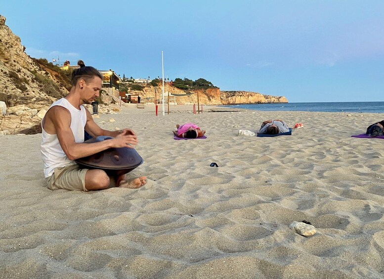 A person sitting on a sandy beach with a drum, a pink towel, and a bag nearby. 1