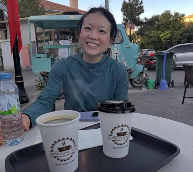 A person seated at a table with two coffee cups and a water bottle. 2