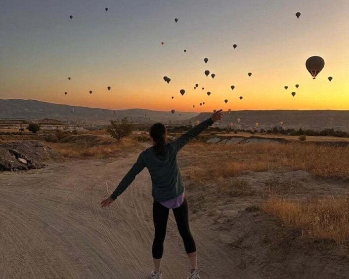 A person standing on a dirt path with arms outstretched, looking at hot air balloons in the sky during sunset. 1