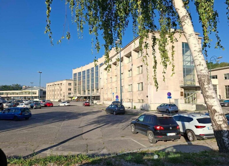 Parkkipaikka, jossa on useita autoja, rakennus suurilla ikkunoilla ja puu, jonka oksista roikkuu lehtiä. 1