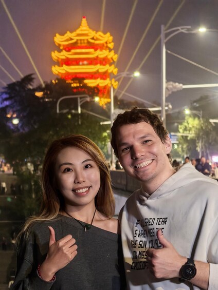 Two people giving thumbs up in front of a brightly lit pagoda at night. 3