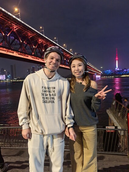 Two people standing in front of a bridge with a city skyline and a tower in the background. 5