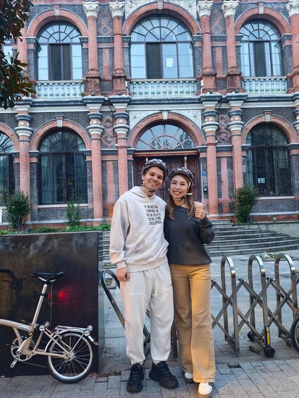 Two people standing in front of a historic building with arched windows and decorative brickwork. 4