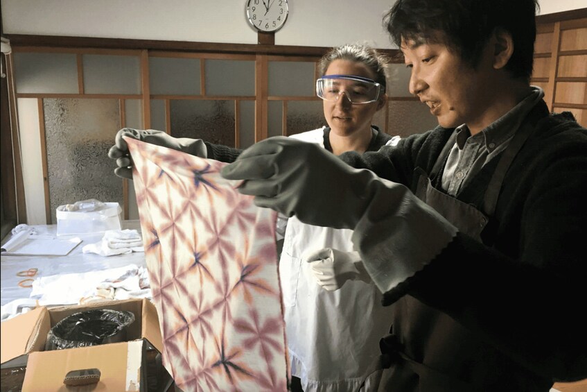Two people examining a patterned fabric in a room with wooden paneling and a clock on the wall. 1