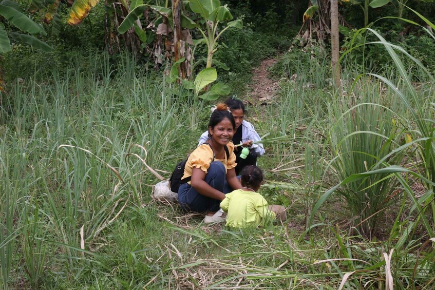 Two people sitting on the ground in a grassy area with tall plants and trees in the background. 1