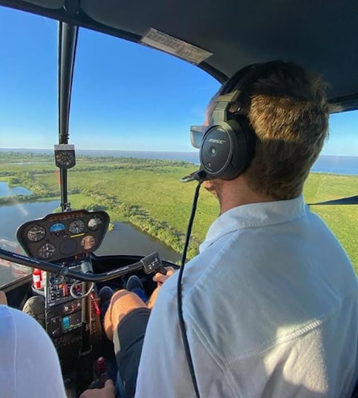 Henkilö helikopterin ohjaamossa, josta avautuu näkymä alla olevaan vihreään maisemaan. 2
