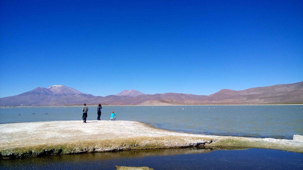 A group of people standing on a concrete platform by a calm lake with mountains in the background. 2
