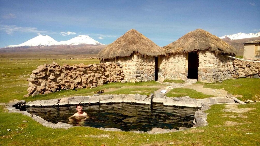 A stone structure with thatched roofs, a stone wall, and a small pond in a grassy field with mountains in the background. 5
