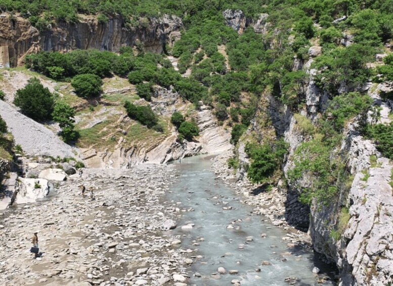 A rocky riverbed with a person standing on the left, surrounded by steep cliffs and dense greenery. 2