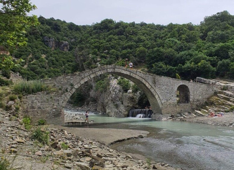 An ancient stone bridge over a river with a waterfall, surrounded by lush greenery. 4
