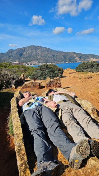 Two people lying on a stone bench with a coastal landscape and mountains in the background. 2