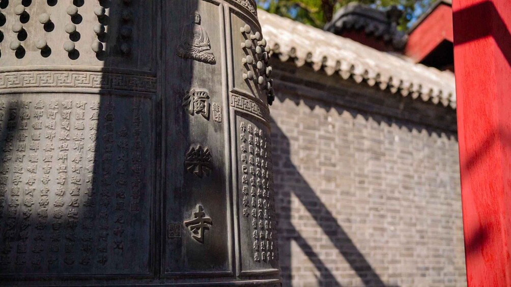 A traditional Chinese bell with inscriptions, set against a brick wall with a tiled roof. 1