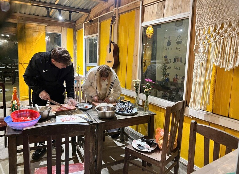 Two people preparing food at a wooden table with yellow walls, a guitar, and a chalkboard with writing. 1