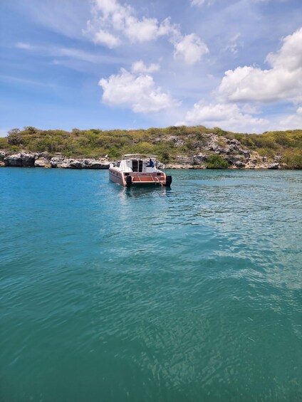 A boat named "CHERICO" on a turquoise sea with a rocky shoreline and sparse vegetation in the background. 4