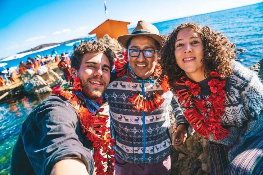 Three people with red flower garlands, a patterned sweater, and a hat, standing by a body of water with a clear sky. 4