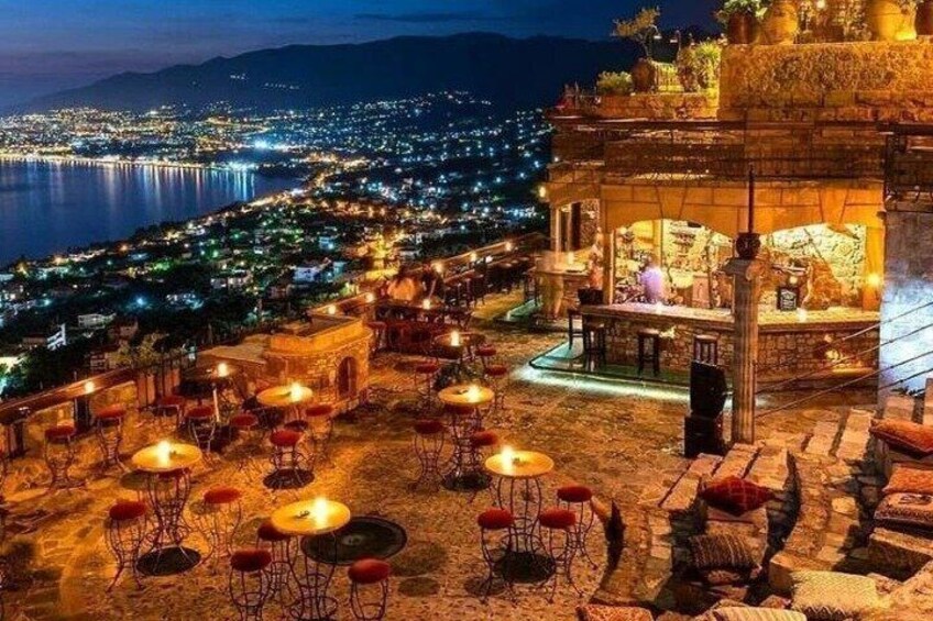 A terrace with tables and chairs, overlooking a cityscape and a body of water at night. 1
