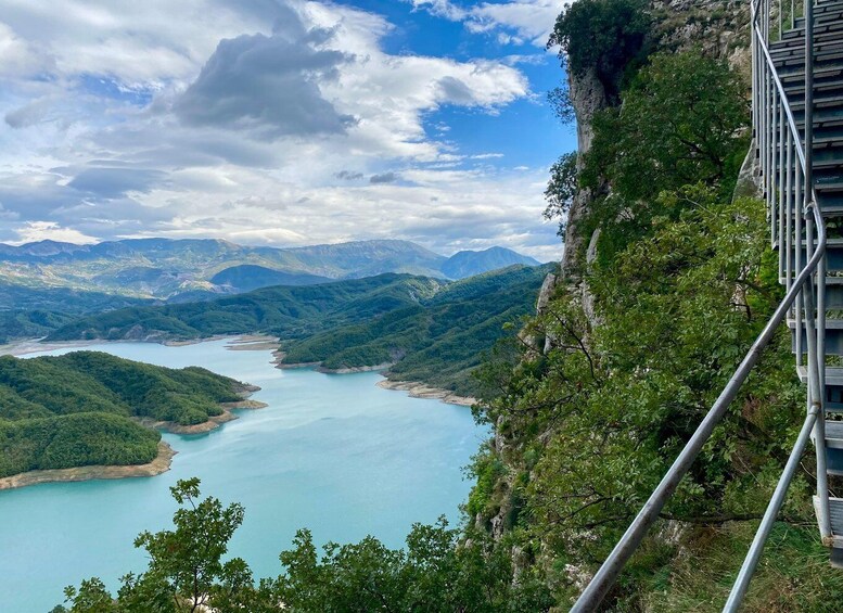 A mountainous landscape with a clear blue lake, a metal bridge, and lush greenery. 3