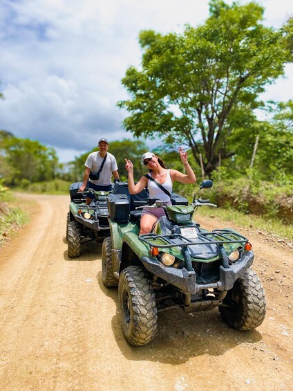Two people on ATVs on a dirt road with trees in the background. 1