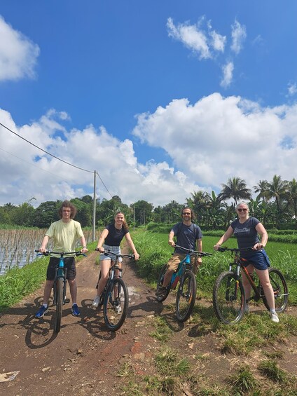 Three people on bicycles on a rural dirt path with a water body and palm trees in the background. 3