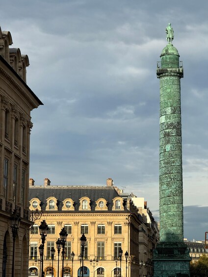 A tall, green monument with a statue on top, surrounded by historic buildings with ornate windows and street lamps. 4