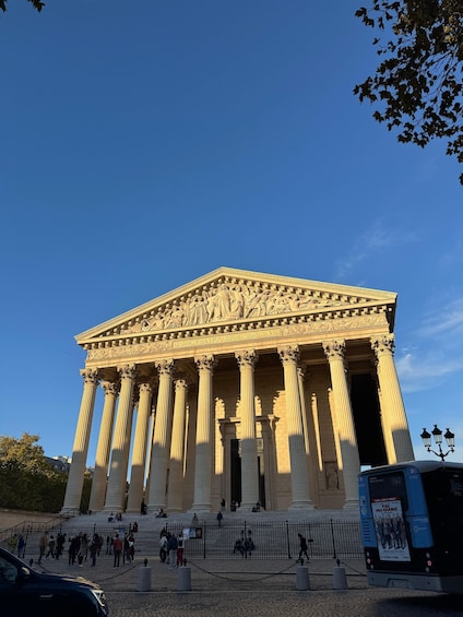 A classical building with columns and detailed carvings, surrounded by a paved area with people and a bus. 3