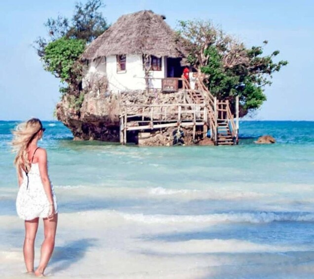 A woman standing on a beach, a thatched-roof house on stilts, and clear turquoise water. 4