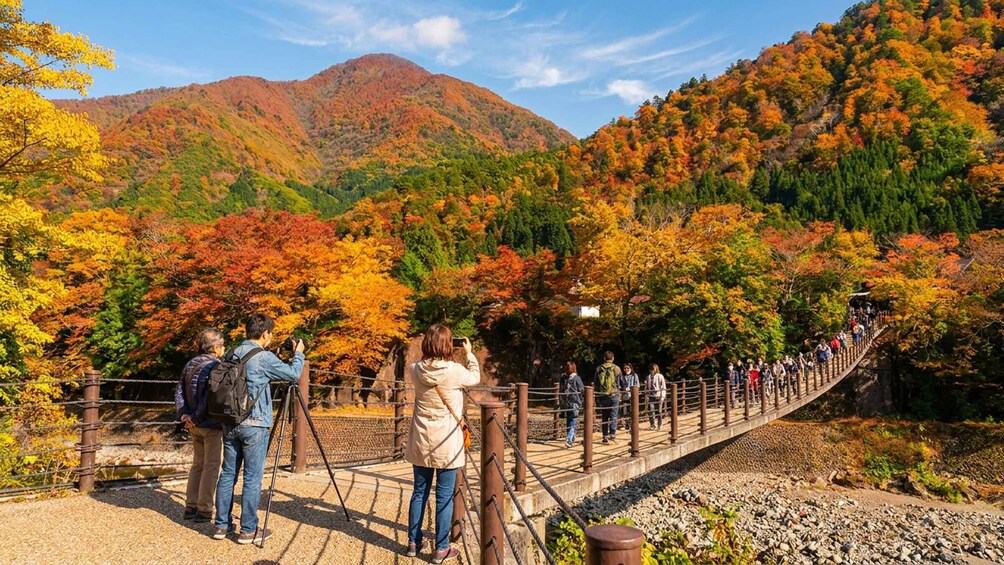 A wooden bridge with people taking photos, surrounded by autumn-colored trees and mountains. 4