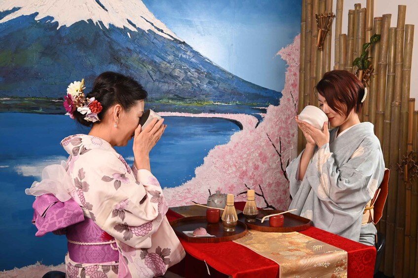 Two people in traditional attire at a table with a tea ceremony setup, including a teapot, cups, and a bamboo whisk, against a backdrop of a painted landscape with cherry blossoms and a mountain. 1