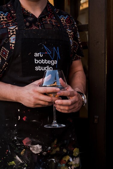 A person holding a glass with a drink, wearing a patterned shirt and an apron with 'aru bottega studio' text. 2
