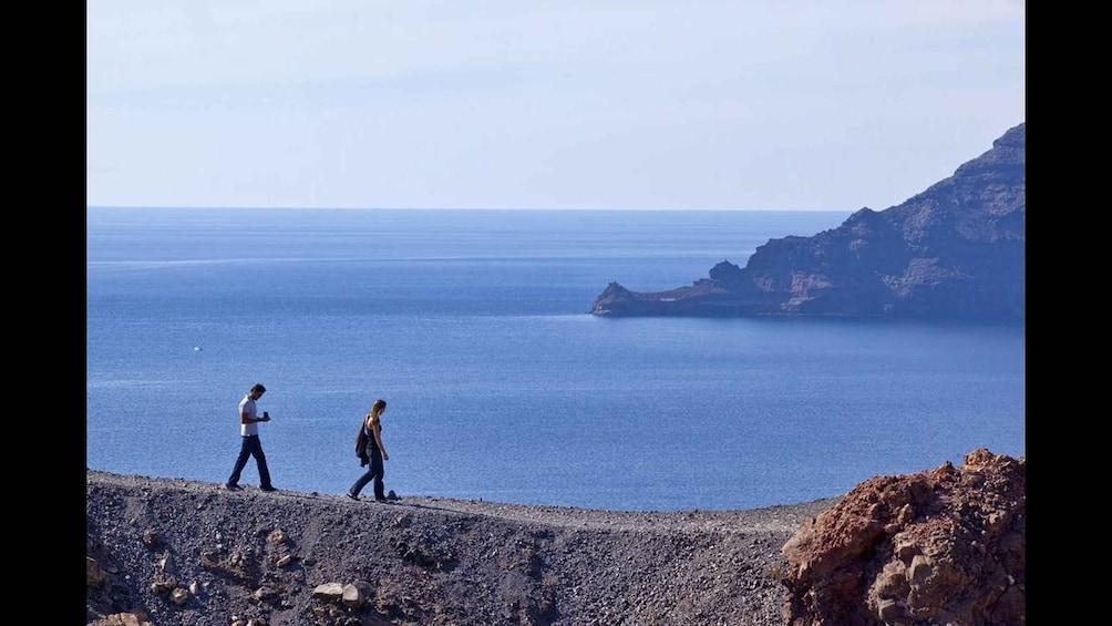 Two people walking along a rocky shoreline with a calm sea and cliffs in the background. 4