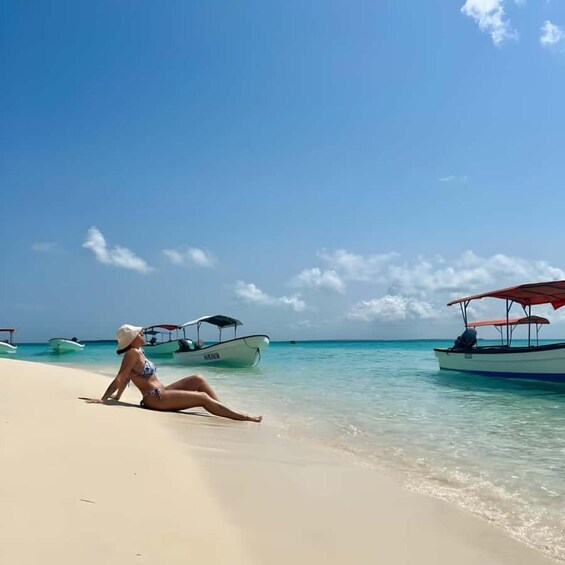 A woman sitting on a sandy beach, boats in the water, and a clear blue sky. 1