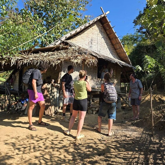 A group of people standing in front of a rustic thatched-roof hut with a dirt ground and trees in the background. 1