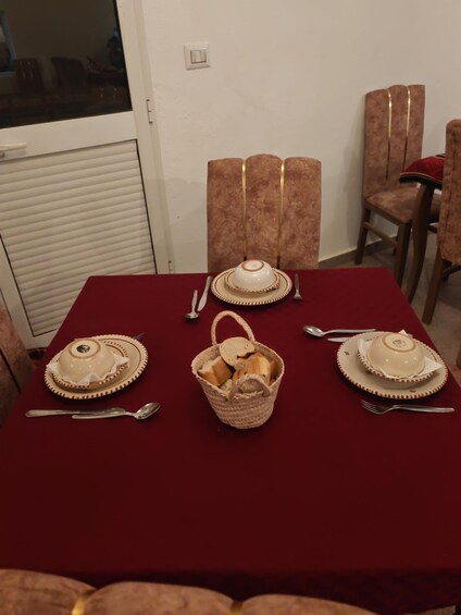 A dining table with a red tablecloth, two white ceramic serving dishes, a woven basket with bread, and a window with blinds. 5