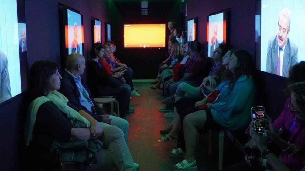 A group of people seated in a dimly lit room with large screens displaying images. 4