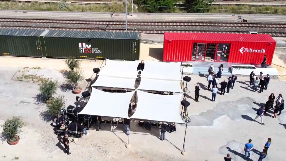 A train station with a red building, a green container, and a white tent with people around. 5