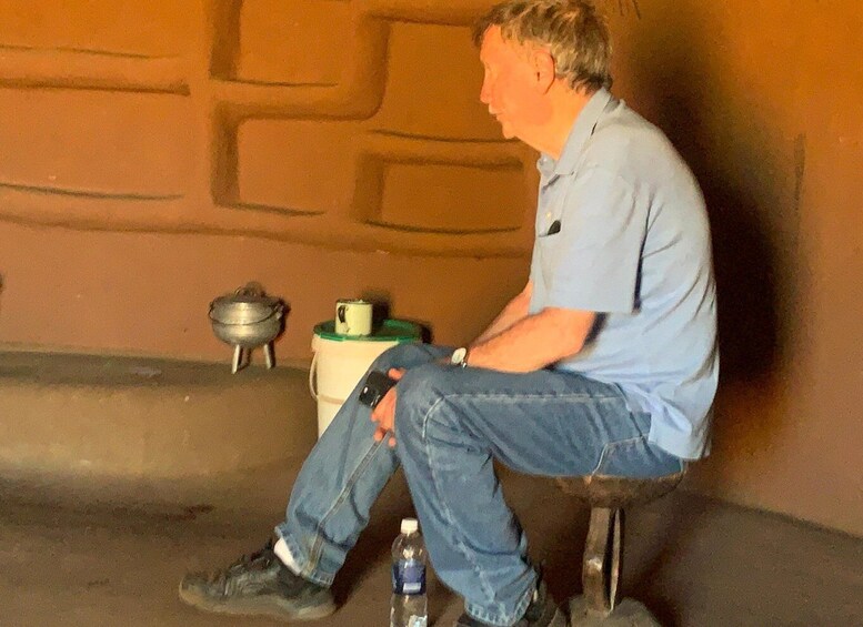 A man sitting on a stool with a water bottle beside him, a pot, and a mug on a table in the background. 2