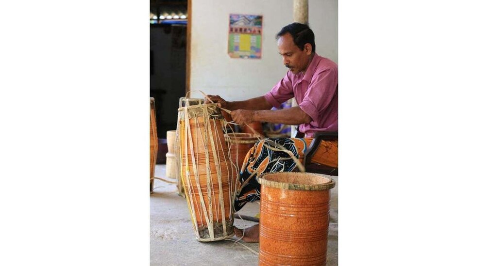 A person seated, working on a large, vertically oriented, wooden instrument with strings, possibly a traditional stringed instrument, in a room with a concrete floor and a poster on the wall. 5