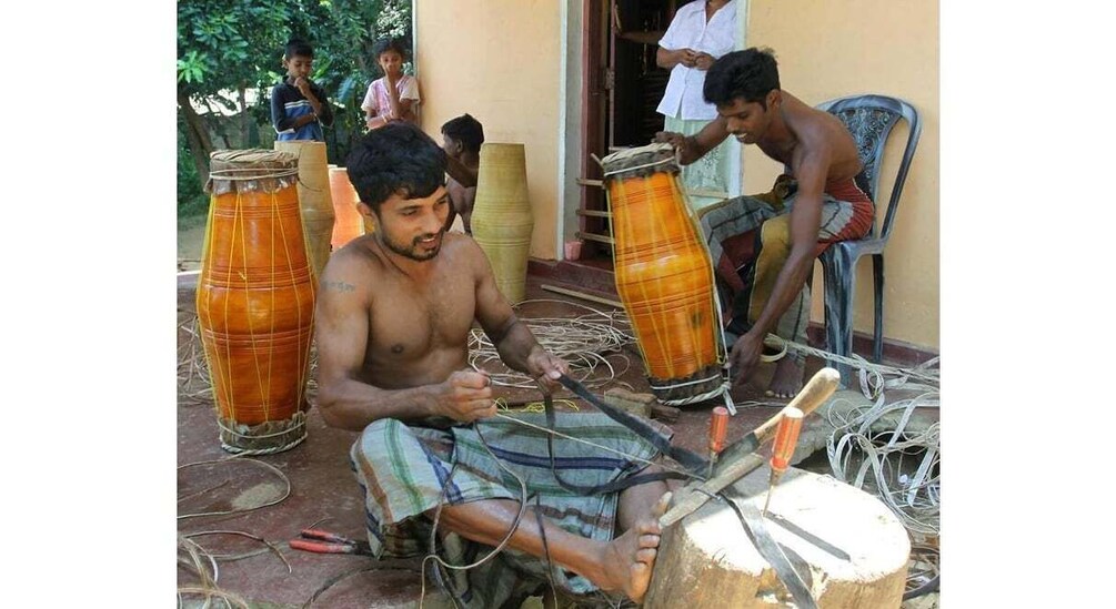 A man seated outdoors, working on a drum with tools and wires around him. 1