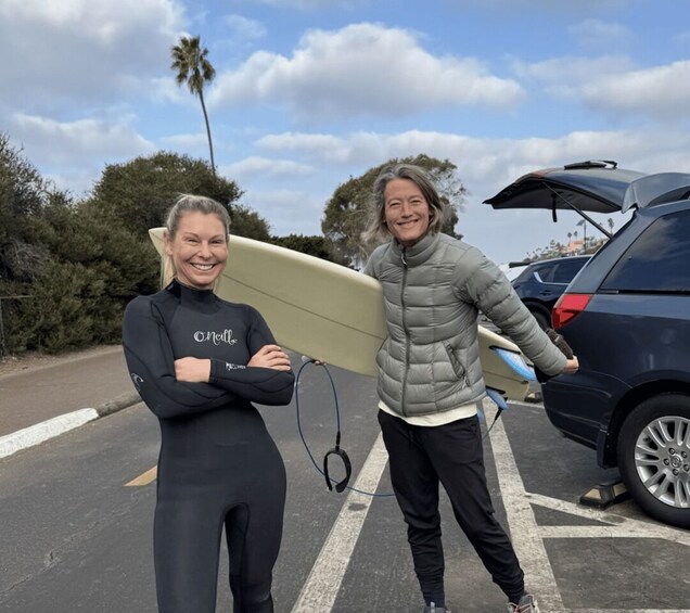 Two people standing in a parking lot with surfboards, one holding a surfboard and the other with a car's trunk open. 2
