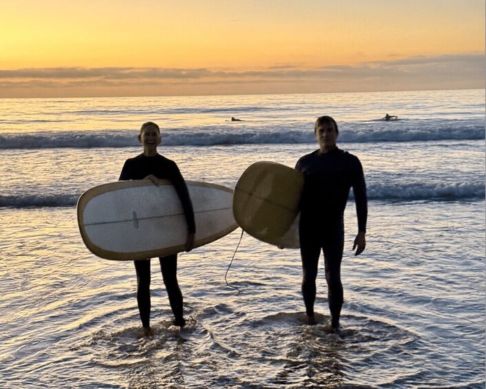 Two people standing in shallow water at the beach, each holding a surfboard. 4