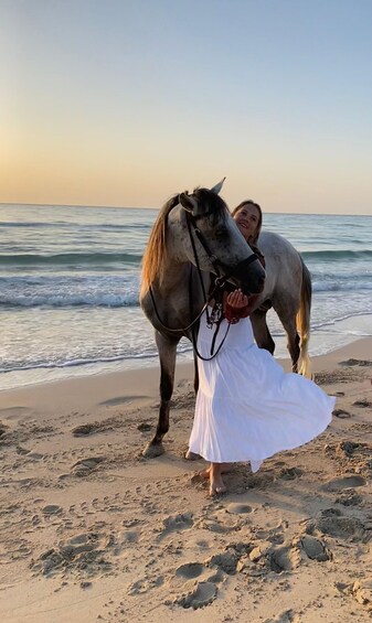 A person in a white dress standing on a beach with a horse. 2