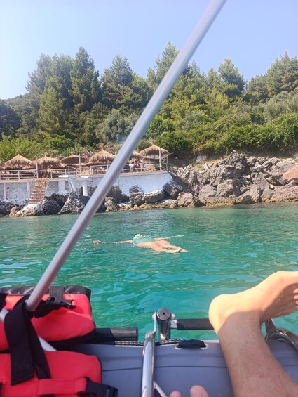 Eine Person schwimmt in klarem Wasser vor einer felsigen Küste und Gebäuden mit Strohdächern. 4