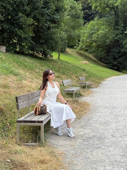 A woman in a white dress sitting on a wooden bench in a park with trees and a gravel path. 3
