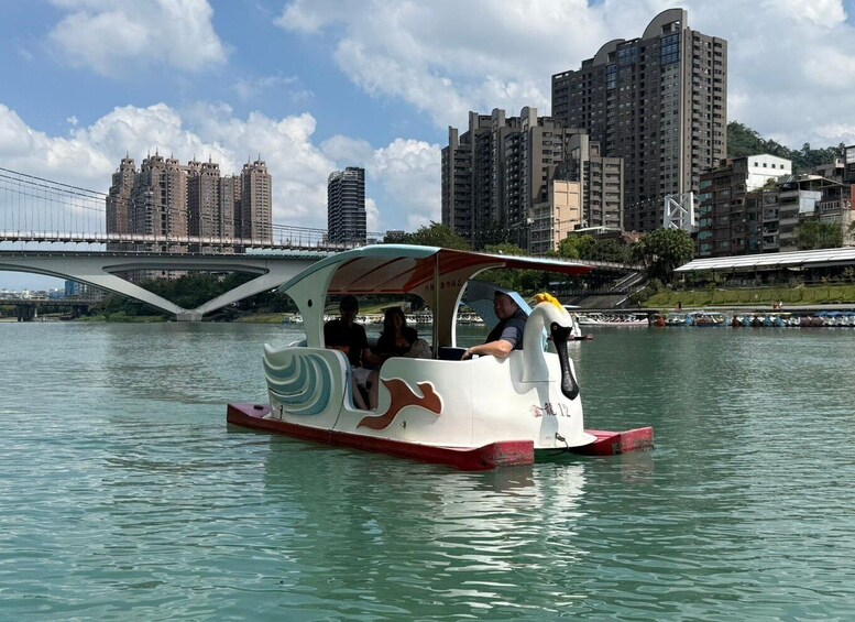 A swan-shaped boat with passengers on a river, a bridge, and high-rise buildings in the background. 5