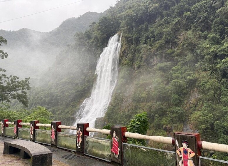 A waterfall cascading down a lush green hillside, with a viewing platform and decorative barriers in the foreground. 1