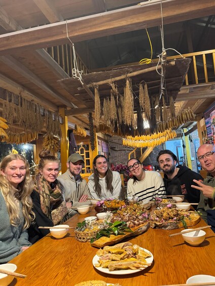 A group of people seated around a wooden table with various dishes and hanging dried goods in a rustic setting. 5