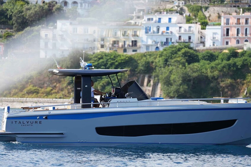 A yacht named 'ITALYURE' with a blue stripe, docked near a coastal town with buildings and greenery. 1