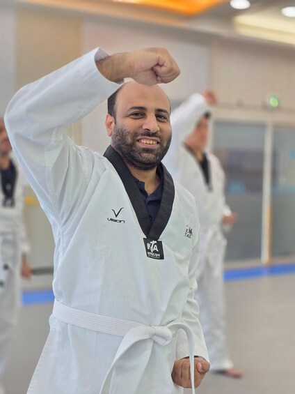 A person in a white martial arts uniform with a black belt and a patch, in a training hall with other individuals in similar attire. 1